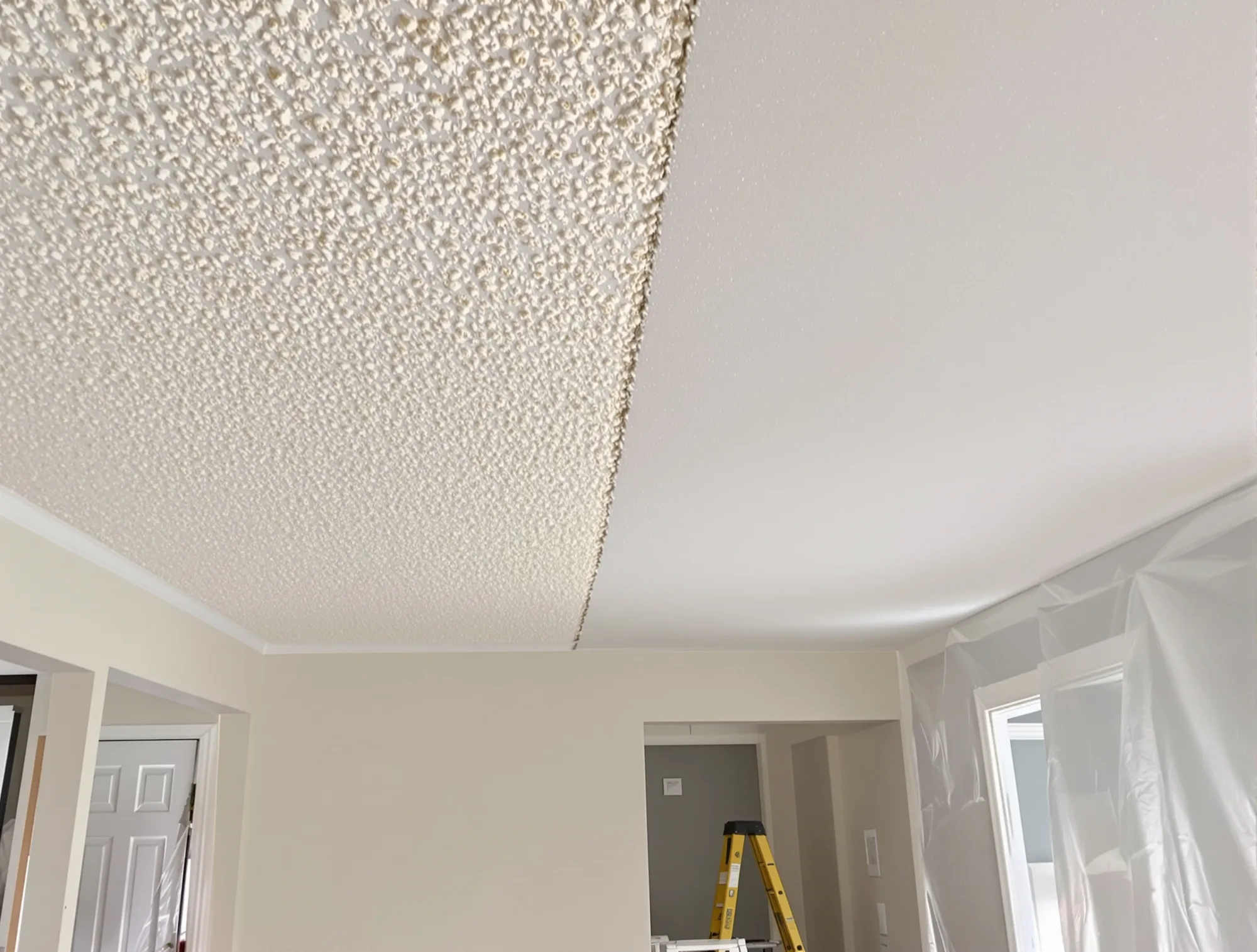 Living room ceiling half scraped smooth and half still covered in popcorn texture, with the room wrapped in plastic